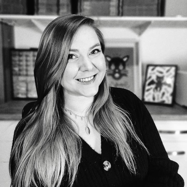 Black and white portrait of Amanda Kinast, owner of Scaredy Cat Studio. She sitting in her office, facing the camera and smiling. 